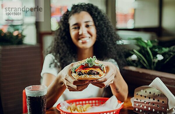 Glückliches Mädchen zeigt Burger in einem Restaurant. Nahaufnahme eines Mädchens  das einen appetitlichen Hamburger in einem Restaurant zeigt