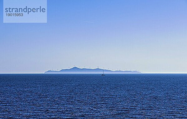 Seelandschaft von ruhigen azurblauen Meer und blauen Himmel mit keine Wolken. Kleines Segelboot und entfernte Insel in dunstigen Hintergrund mit Silhouetten von elektrischen Turbine Windmühlen punktiert. Mittelmeer  Griechenland  Europa