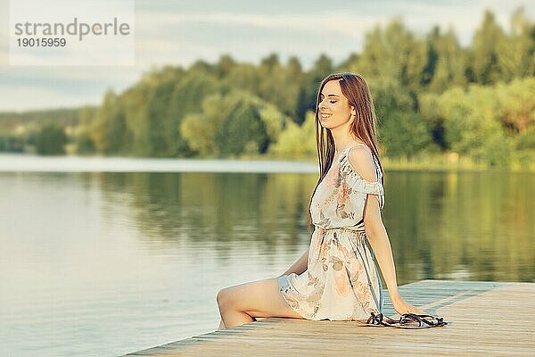 Junge lächelnde Frau entspannt sich mit geschlossenen Augen auf dem Pier am Fluss