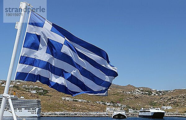 Die griechische Flagge weht vor der typischen griechischen Insellandschaft. Im Hintergrund liegen Kreuzfahrtschiffe und Fähren vor Anker. Heller Sommertag. Inselhüpfen  Abenteuer und Erkundung des Mittelmeers. Selektiver Fokus