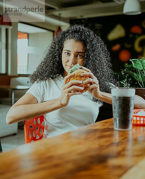 Schönes lateinisches Mädchen  das einen leckeren Hamburger in einem Restaurant genießt. Porträt eines Afro Mädchens  das einen Hamburger in einem Restaurant hält