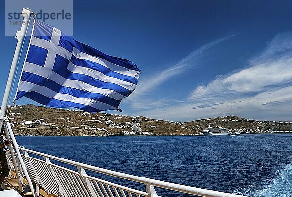 Die griechische Flagge weht vor der typischen griechischen Insellandschaft. Im Hintergrund liegen Kreuzfahrtschiffe und Fähren vor Anker. Heller Sommertag. Inselhüpfen  Abenteuer und Erkundung des Mittelmeers. Selektiver Fokus