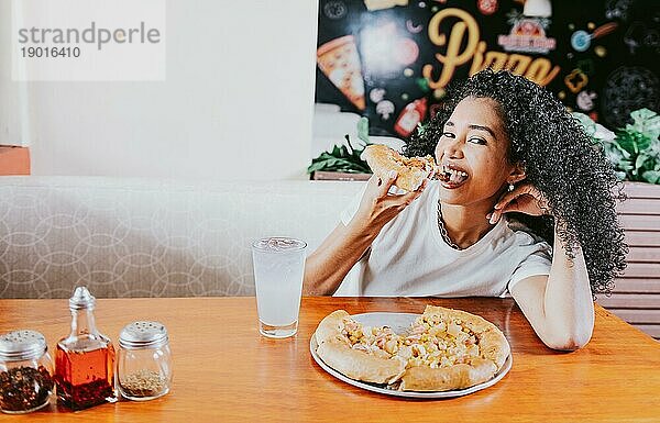 Lächelnde junge Frau  die eine Pizza in einem Restaurant genießt. Glückliches afrohaariges Mädchen isst Pizza in einem Restaurant  isst und schaut in die Kamera