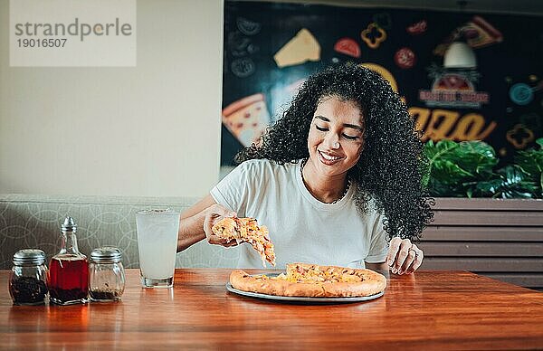 Glückliche afrohaarige Frau isst Pizza in einem Restaurant. Lebensstil der afrohaarigen Frau  die eine Pizza in einem Restaurant genießt