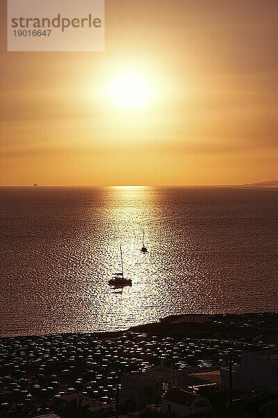 Wunderschöne Meereslandschaft bei Sonnenuntergang. Niedrige Sonne in dunstigen Himmel und Sonne Blendung auf ruhigem Meer Wasser. Silhouetten von kleinen Segelbooten gehen auf das Meer in Sonnenlicht Streifen  dunkle Küste im Vordergrund. Sommer heiße Tage  Urlaub  Freizeit  romantisches Segeln. Vertikale Aufnahme