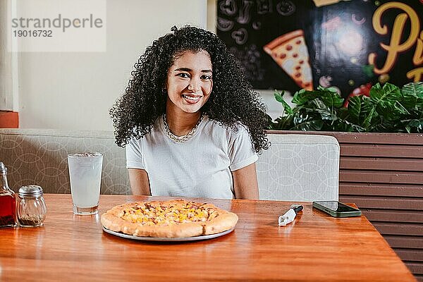 Lächelnd Junge lockige Frau sitzt in einer Pizzeria. Porträt eines schönen Mädchens in einer Pizzeria