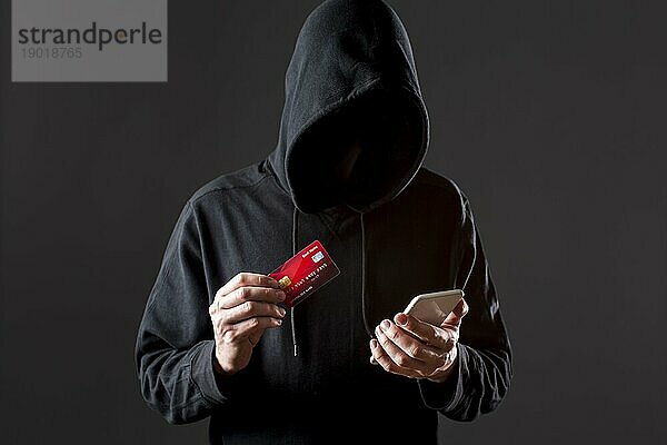 Frontansicht männlicher Hacker  der eine Smartphonekreditkarte hält. Foto mit hoher Auflösung