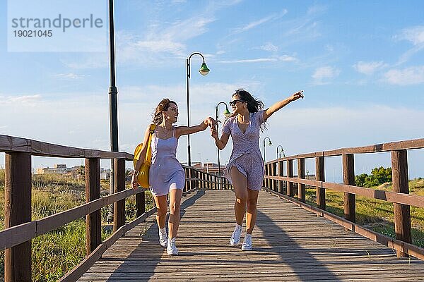 Freundinnen im Sommerurlaub am Strand  die auf einem Holzpfad spazieren gehen und vor Aufregung springen
