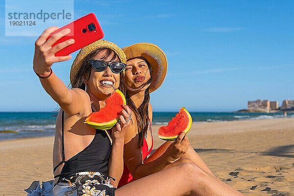 Freunde am Strand im Sommer essen eine Wassermelone mit dem Meer im Hintergrund und machen ein Selfie mit dem Handy