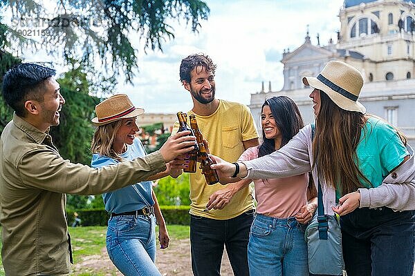 Eine multiethnische Gruppe von Freunden feiert in einem Stadtpark mit Bier. Lächelnd und mit den Flaschen anstoßend