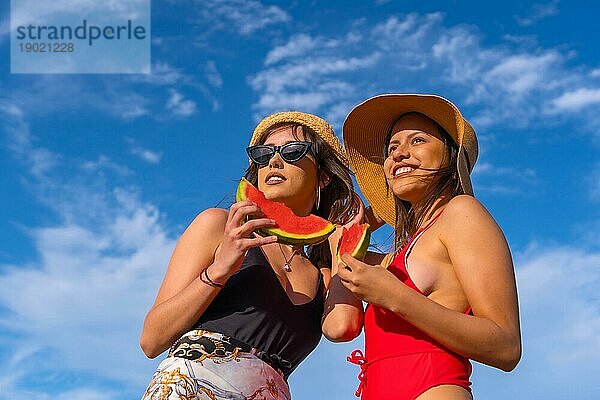 Porträt von Freundinnen am Strand im Sommer lächelnd essen eine Wassermelone mit dem Himmel im Hintergrund  von unten nach oben Schuss