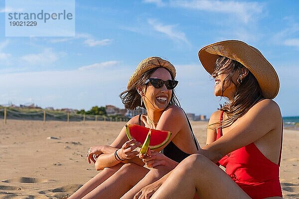 Porträt von Freundinnen im Sommerstrandurlaub  die eine Wassermelone essen  mit dem Meer im Hintergrund  auf dem Sand sitzend