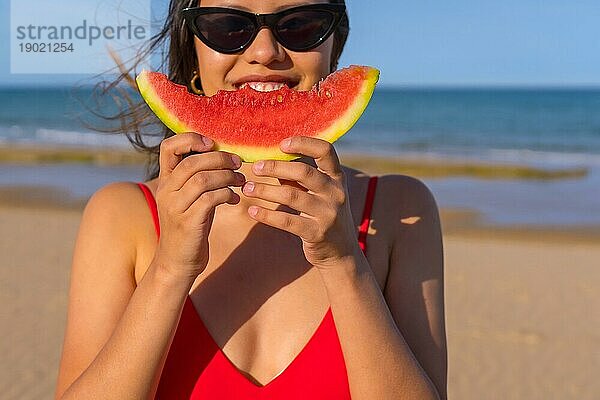 Porträt einer Frau  die am Strand eine Wassermelone isst. Nahaufnahme lächelnd mit Bikini