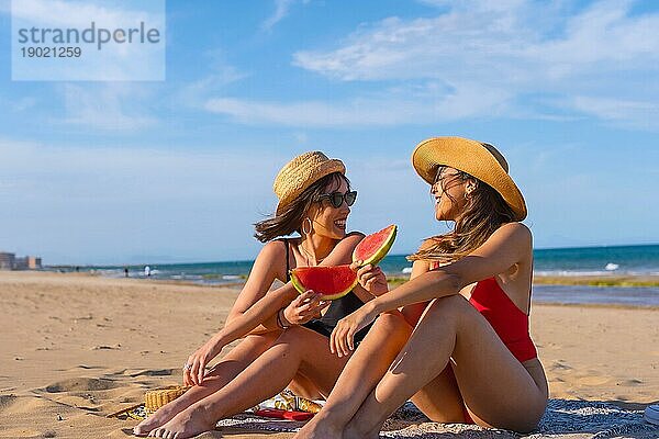 Weibliche Freunde im Urlaub am Strand im Sommer essen eine Wassermelone mit dem Meer im Hintergrund  sitzen auf dem Sand