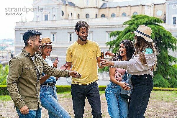 Eine multiethnische Gruppe von Freunden feiert in einem Stadtpark mit Bier. Sommervergnügen  Grüße an den neuen Freund  der angekommen ist