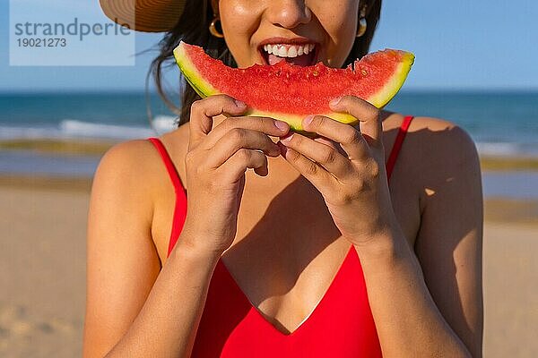 Porträt einer Frau  die am Strand eine Wassermelone isst. Ausschnitt eines lächelnden Mundes mit rotem Bikini