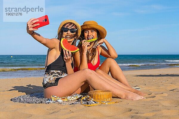 Freunde am Strand im Sommer essen eine Wassermelone mit dem Meer im Hintergrund und machen ein Selfie