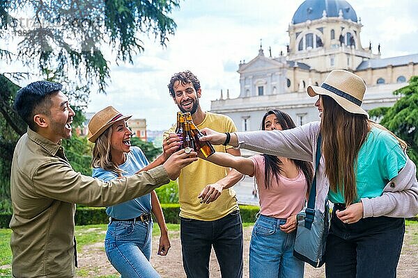 Eine multiethnische Gruppe von Freunden feiert in einem Stadtpark mit Bier. Lächelnd und mit den Flaschen anstoßend