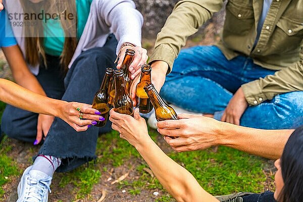Eine Gruppe multiethnischer Freunde sitzt im Stadtpark und unterhält sich mit Bierflaschen neben einem Baum