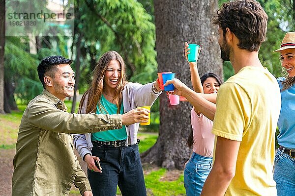 Multiethnische Gruppe von Geburtstagsgästen im Stadtpark  die mit bunten Bechern anstoßen