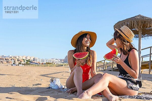 Freundinnen im Urlaub am Strand im Sommer essen eine Wassermelone  sitzen auf dem Sand neben einem Sonnenschirm