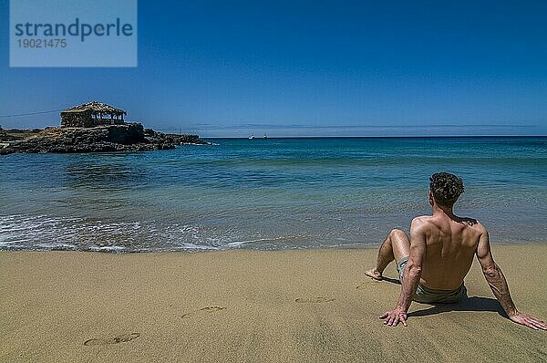 Junger Mann mit Badehose  sitzend am Sandstrand. Tarrafal. Santiago. Cabo Verde. Afrika