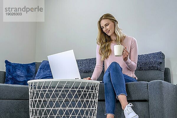 Nette schöne Frau mit blonden Haaren arbeitet am Laptop  während sie sich zu Hause auf das Sofa setzt