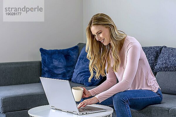 Nette schöne Frau mit blonden Haaren arbeitet am Laptop  während sie sich zu Hause auf das Sofa setzt
