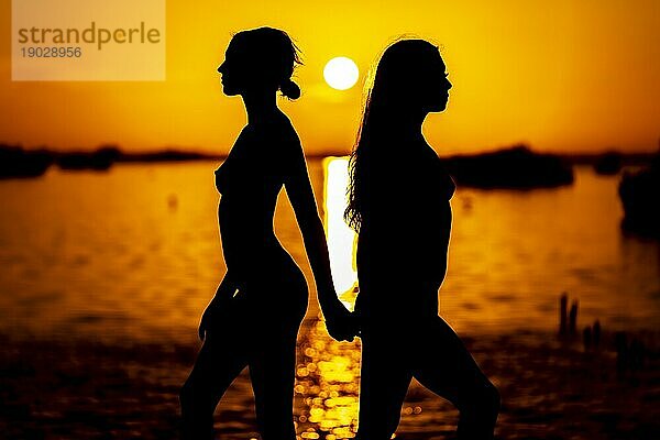 Zwei schöne nackte lateinische Modelle sind gegen die aufgehende Sonne hinter ihnen auf einem exotischen karibischen Strand silhouetted