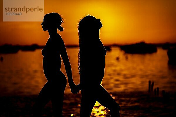 Zwei schöne nackte lateinische Modelle sind gegen die aufgehende Sonne hinter ihnen auf einem exotischen karibischen Strand silhouetted
