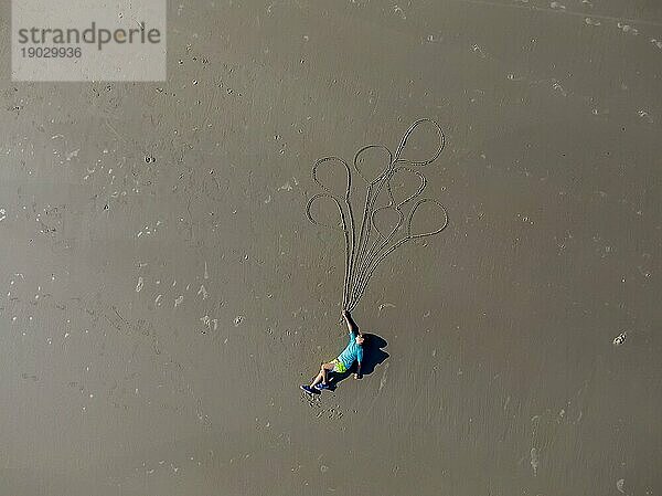 Ein kleines Kind tut so  als würde es im Urlaub am Strand mit Luftballons davongetragen