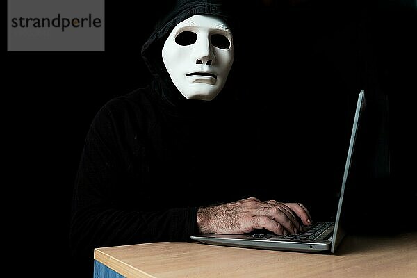 Schwarz gekleideter Hacker mit Maske und Kapuze tippt auf einem Computer  während er in die Kamera schaut CybercrimeKonzept