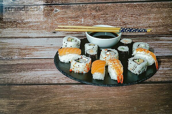 Sushisortiment auf einer schwarzen Schieferplatte mit Sauce und Stäbchen auf einem Holztisch