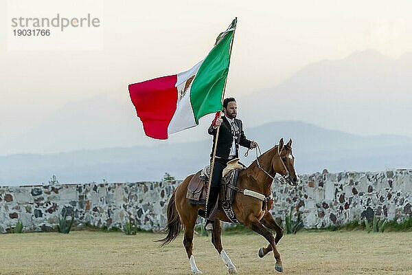 Ein stattlicher mexikanischer Charro posiert vor einer Hazienda in der mexikanischen Landschaft und hält die mexikanische Flagge