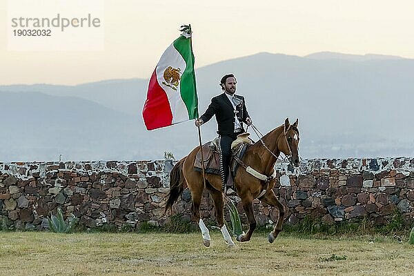 Ein stattlicher mexikanischer Charro posiert vor einer Hazienda in der mexikanischen Landschaft und hält die mexikanische Flagge