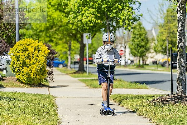 Ein Kind fährt mit seinem Roller auf einem Bürgersteig in einem Vorort und trägt eine Schutzmaske  um sich vor Viren zu schützen