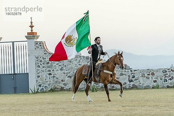 Ein stattlicher mexikanischer Charro posiert vor einer Hazienda in der mexikanischen Landschaft und hält die mexikanische Flagge