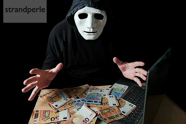 Hacker Mann in schwarz gekleidet mit weißer Maske und schwarzer Kapuze mit einem Stapel von Eurobanknoten auf seinem Laptop CybercrimeKonzept vor schwarzem Hintergrund