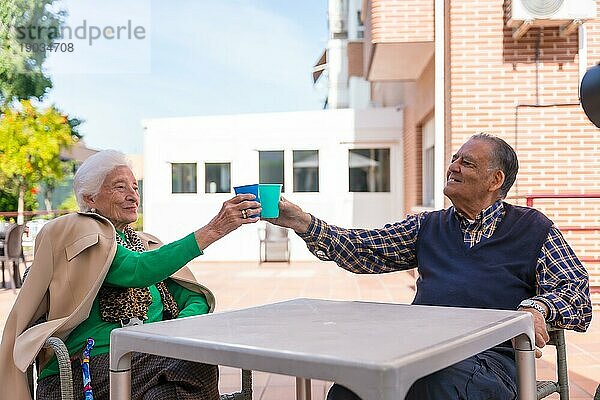 Zwei ältere Menschen im Garten eines Pflegeheims  die an einem sonnigen Sommertag mit leuchtenden Gläsern anstoßen