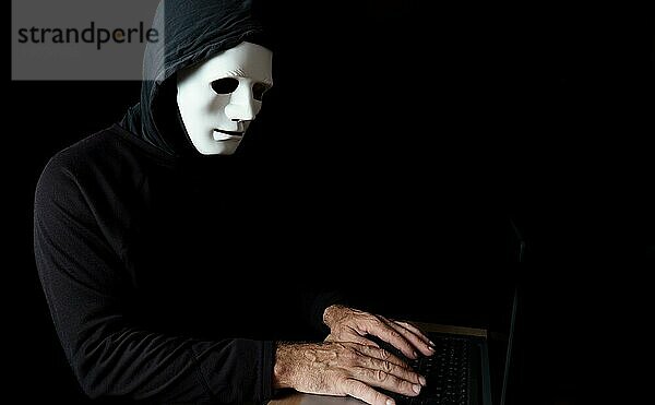 Männlicher Hacker  schwarz gekleidet mit Kapuze und weißer Maske  der einen Computer benutzt