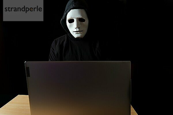 Männlicher Hacker in schwarzer Kleidung mit Kapuze und weißer Maske hinter einem Computer