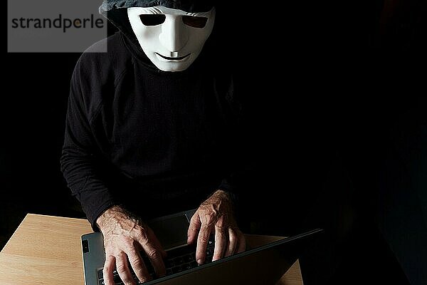 Schwarz gekleideter Hacker mit Kapuze und Maske tippt auf einem Computer CybercrimeKonzept