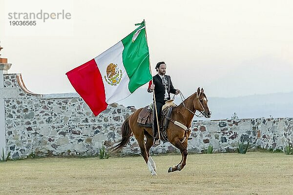 Ein stattlicher mexikanischer Charro posiert vor einer Hazienda in der mexikanischen Landschaft und hält die mexikanische Flagge