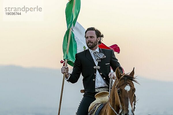 Ein stattlicher mexikanischer Charro posiert vor einer Hazienda in der mexikanischen Landschaft und hält die mexikanische Flagge