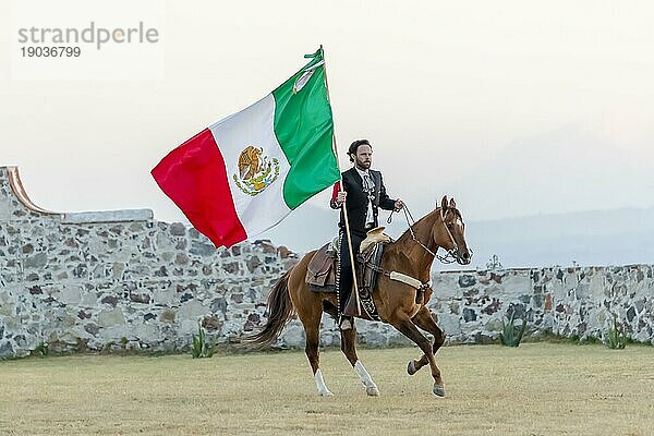 Ein stattlicher mexikanischer Charro posiert vor einer Hazienda in der mexikanischen Landschaft und hält die mexikanische Flagge