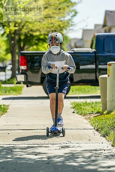 Ein Kind fährt mit seinem Roller auf einem Bürgersteig in einem Vorort und trägt eine Schutzmaske  um sich vor Viren zu schützen