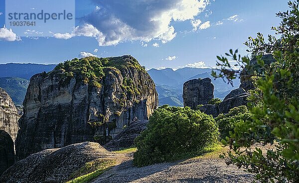 Abendansicht der typischen Meteora Landschaft in Griechenland. Massive Sedimentgesteine  einzelne Felsen  ferne Berge und ein Tal dazwischen. Blauer Himmel und große Wolken. Selektiver Fokus. Busch im Vordergrund