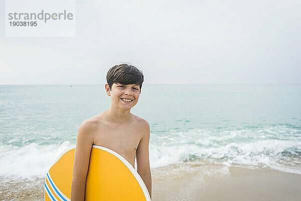 Vorderansicht eines jungen Teenagers  der am Strand steht und ein Surfbrett hält