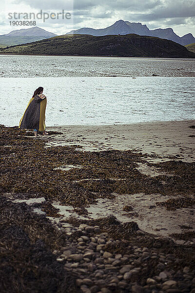 Frau im langen Mantel spaziert am Strand in Schottland