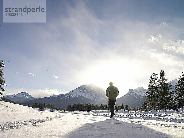 Mann geht die schneebedeckte Straße entlang in Richtung Sonne und Berge